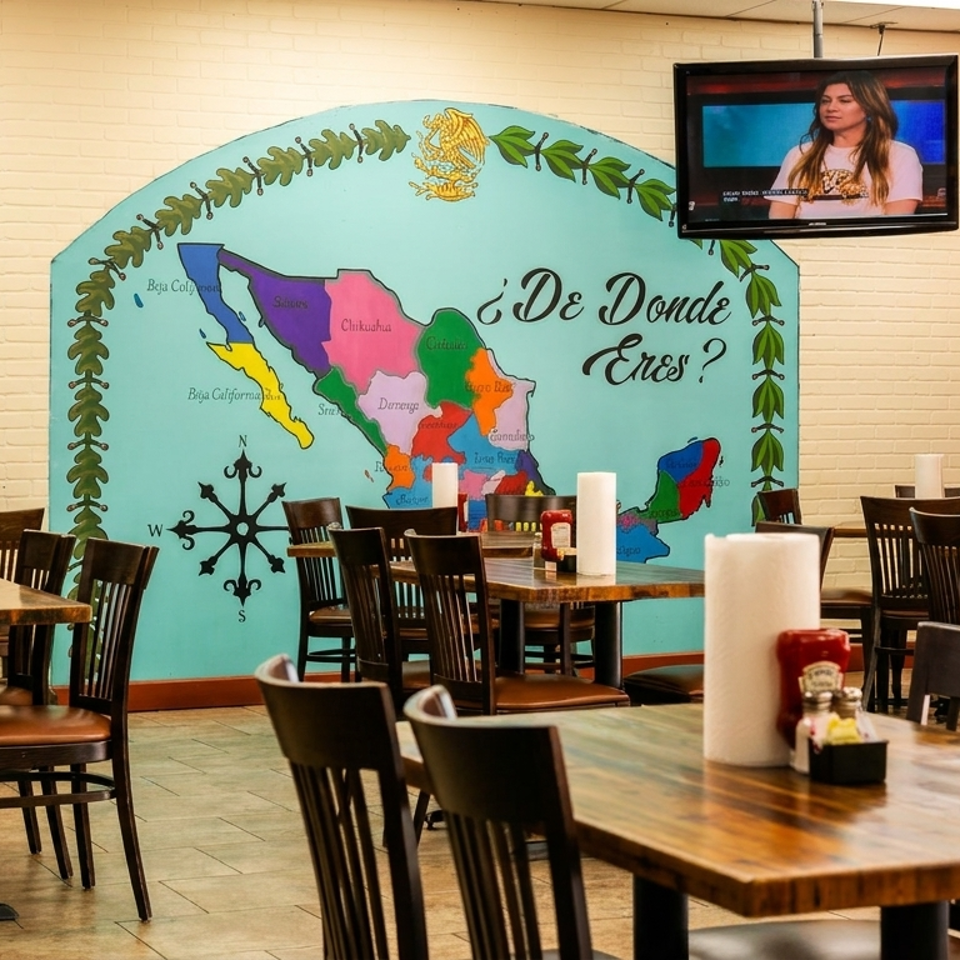 Empty restaurant dining room with wooden tables, chairs, condiments, and a colorful Mexico map mural beneath a television.